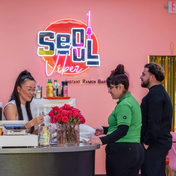 A Seoul Vibez employee helps customers at the counter, with a neon sign glowing behind them.
