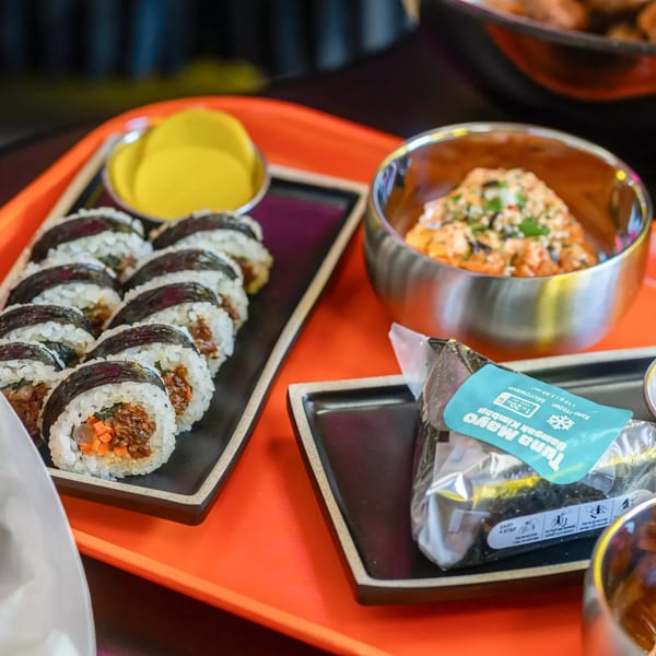 Plates of kimbap rolls, a bowl of kimchi, and a packaged tuna mayo samgak kimbap on an orange tray.