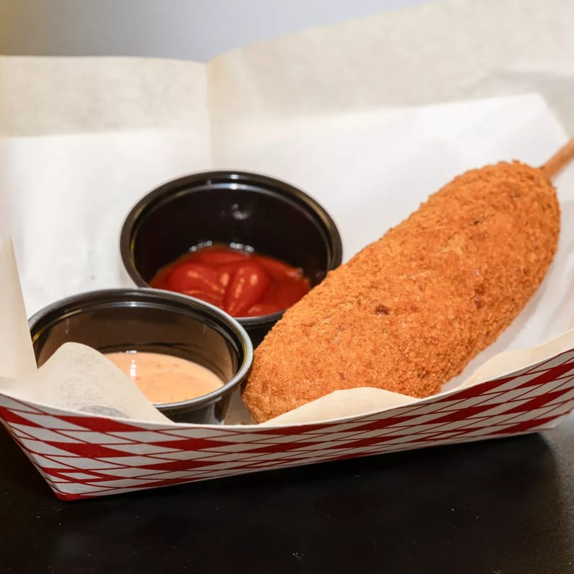 A crispy Korean corn dog with ketchup and a creamy sauce in a red and white striped tray.