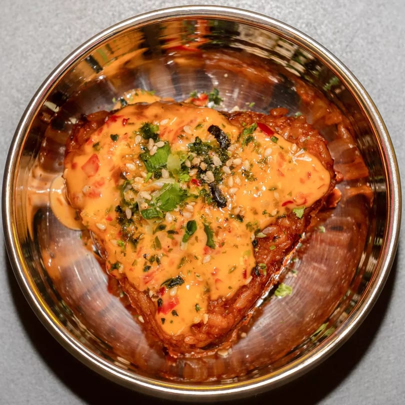 Crispy fried chicken topped with spicy orange sauce, sesame seeds, and herbs in a metal bowl.